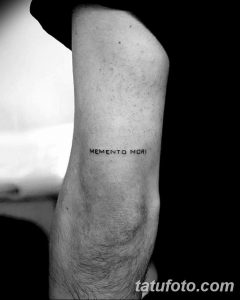 Фото минималистичной татуировки с надписью 'Memento Mori' на локте. Тонкие линии, простой шрифт.