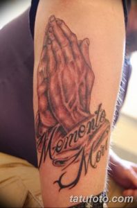 Фото татуировки на руке с изображением молящихся рук и надписи 'Memento Mori'. Реалистичный рисунок с использованием теней и полутонов.