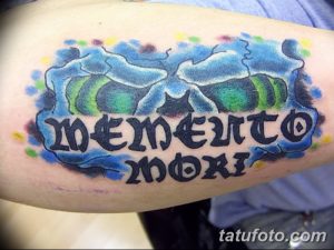 Фото тату надписи 'Memento Mori' в акварельной технике на руке. Использованы синие, зеленые и желтые оттенки, создающие эффект размытости.