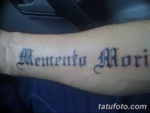 Фото тату с надписью 'Memento Mori' в готическом стиле на предплечье. Черно-серая татуировка с детализированным шрифтом.