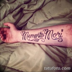 Фото тату надписи 'Memento Mori' изящным, витиеватым шрифтом на предплечье. Черный рисунок татуировки выглядит элегантно и женственно.
