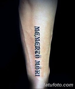 Фото тату надписи 'Memento Mori' готическим шрифтом на внутренней стороне предплечья. Черный рисунок татуировки четкий и контрастный.