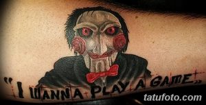 Фото тату на руке с изображением Джокера из фильма "Пила" и цитатой "I wanna play a game". Детализированный рисунок, яркие цвета, мрачная атмосфера.