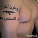 Фото тату: автомат Калашникова с арабской надписью на плече мужчины. Черно-белый рисунок татуировки в реалистичном стиле.