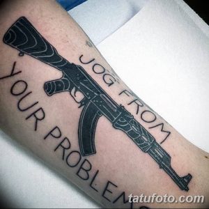 Фото татуировки на руке, изображающей автомат Калашникова и надпись 'JOG FROM YOUR PROBLEM'. Черно-белый рисунок в реалистичном стиле.