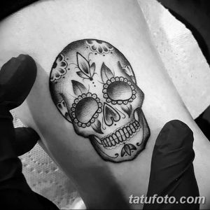 Фотография красочной татуировки в стиле сахарной черепушки (sugar skull) на руке. Детализированный рисунок с использованием черного и серого цветов, украшенный цветочными узорами.