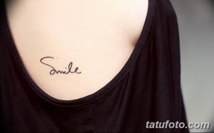 Фото тату: надпись "Smile" каллиграфическим шрифтом на груди. Минималистичный рисунок татуировки, выполненный черными чернилами.