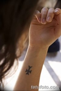 Фото тату: небольшой черный крест на запястье девушки. Простой и лаконичный дизайн, выполненный в минималистичном стиле.