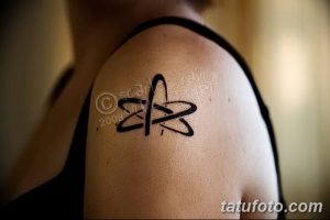 Фотография минималистичной татуировки на плече, изображающей схематичное изображение атома, выполненное черными линиями.