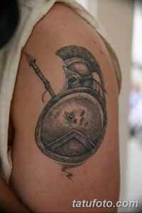 Фото татуировки на плече: изображение древнегреческого щита и копья, выполненное в серой гамме. Стильный и мужественный рисунок.