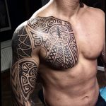Фотография мужчины с обширной татуировкой в стиле маори, покрывающей грудь, плечи и руки. Сложный узор с множеством деталей.