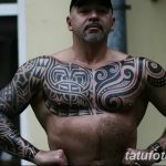 Фотография мужчины с обширной татуировкой в стиле маори, покрывающей грудь, плечи, руки и часть спины.
