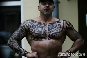 Фотография мужчины с обширной татуировкой в стиле маори, покрывающей грудь, плечи, руки и часть спины.