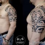 Фото тату: мужское плечо, украшенное сложной татуировкой в полинезийском стиле. Рисунок выполнен в черном цвете и имеет геометрическую форму.