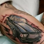 Фотография цветной татуировки на руке, изображающей классический автомобиль. Детализированный рисунок татуировки с реалистичными отражениями.