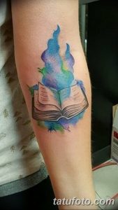 Фото тату на предплечье с изображением горящей книги. Акварельный стиль, яркие синие и фиолетовые оттенки, динамичный и эффектный рисунок.