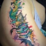 Красочное фото акварельной татуировки на плече с изображением стопки книг, воздушного шара, бабочек и брызг краски. Рисунок выполнен в ярких, пастельных тонах.