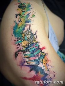 Красочное фото акварельной татуировки на плече с изображением стопки книг, воздушного шара, бабочек и брызг краски. Рисунок выполнен в ярких, пастельных тонах.
