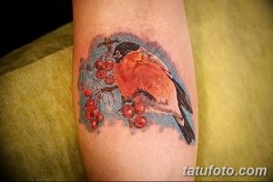 Фото реалистичной татуировки птицы, сидящей на ветке рябины с красными ягодами. Детализированный рисунок татуировки на руке.