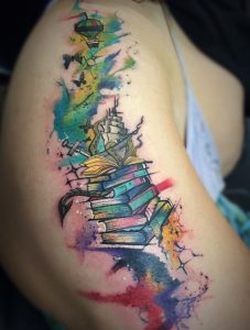 Фото акварельной татуировки на плече с изображением стопки книг, воздушного шара, маяка и брызг краски. Яркие и нежные цвета.