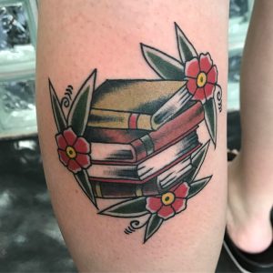 Фото тату на ноге с изображением стопки книг, украшенных красными цветами. Рисунок выполнен в стиле олдскул, с использованием ярких цветов и четких контуров.