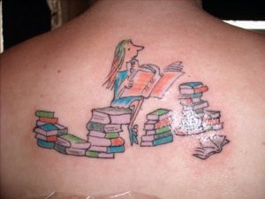 Фото тату на спине с изображением девочки, читающей книгу, сидящей на стопке книг. Рисунок выполнен в ярких, мультяшных тонах.