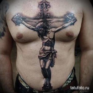 Фото тату: крупное изображение распятия Христа на груди мужчины, выполненное в реалистичном стиле с использованием черных и серых оттенков. Детализированный рисунок татуировки.