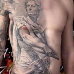 Фото реалистичной татуировки с изображением Юлия Цезаря в доспехах на торсе мужчины. Детализированная прорисовка, использование светотени, эффект объемности.