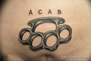 Фото татуировки на груди с изображением кастетов и надписью 'ACAB'. Рисунок выполнен в реалистичном стиле, с использованием черно-серых тонов.