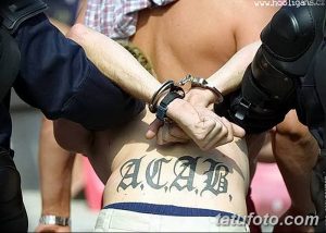 Фото тату надписи 'ACAB' на спине человека, которого задерживает полиция. Черно-белая татуировка, выражающая протест и социальную позицию.