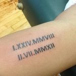 Фото тату: татуировка с римскими цифрами, обозначающими даты 'I.XXIV.MMVIII' и 'II.VII.MMXII' на предплечье. Черно-серый рисунок, четкие линии.