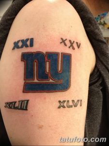 Фото тату на плече: логотип команды New York Giants с римскими цифрами, выполненный в ярких синих и красных цветах.