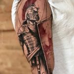 Фото тату на плече: реалистичное изображение римского легионера в доспехах и с щитом, выполненное в черно-красной гамме с высокой детализацией.