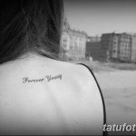 Фото тату с надписью 'Forever Young' выполненной каллиграфическим шрифтом на спине, чуть ниже шеи. Татуировка выполнена в черных тонах.