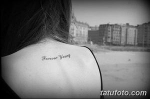 Фото тату с надписью 'Forever Young' выполненной каллиграфическим шрифтом на спине, чуть ниже шеи. Татуировка выполнена в черных тонах.