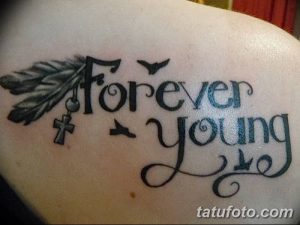 Фото тату 'Forever Young' на плече, украшенное пером, крестом и силуэтами птиц. Рисунок выполнен в черно-серых тонах с использованием теней.