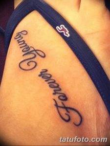 Фото тату с надписью 'Forever Young' выполненной каллиграфическим шрифтом на внутренней стороне предплечья. Татуировка выполнена в черных тонах.