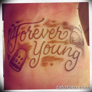 Фото тату с надписью 'Forever Young', украшенной рисунками фотоаппарата и чемодана на мужской груди. Оригинальный дизайн и яркие цвета.