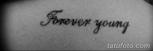 Фото тату с надписью 'Forever Young' черным цветом на коже. Минималистичный дизайн и каллиграфический шрифт.