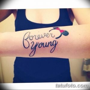 Фото тату с надписью 'Forever Young' и изображением Микки Мауса с бантиком на женском предплечье. Нежная цветовая гамма и игривый дизайн.