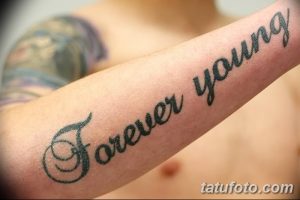 Фото тату с надписью 'Forever Young' черным цветом на мужском предплечье. Изящный шрифт и четкие линии делают эту татуировку стильной и запоминающейся.