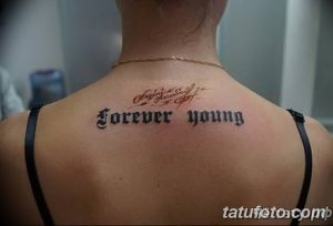 Фотография татуировки с надписью 'Forever Young', выполненной в готическом стиле на спине. Темный и выразительный рисунок татуировки.