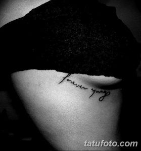 Черно-белое фото татуировки с надписью 'Forever Young' на боку, выполненной каллиграфическим шрифтом. Изящный рисунок татуировки для девушек.