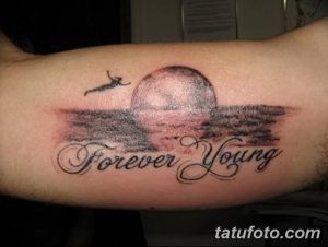 Фото тату надписи 'Forever Young' с изображением заката, моря и летящего самолета. Красивая и символичная татуировка на руке.