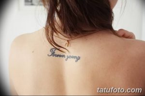Фото тату надписи 'Forever Young' выполненной изящным шрифтом на спине девушки. Нежная и женственная татуировка.