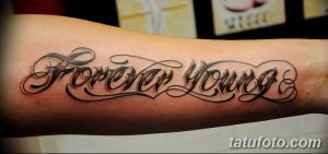Фото тату надписи 'Forever Young' с декоративными завитками и узорами, выполненной в черных тонах на руке. Стильный и привлекательный рисунок.