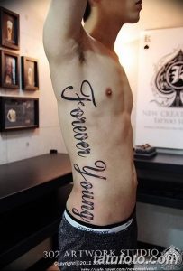 Фото тату с надписью 'Forever Young' на боку мужчины. Большая татуировка, выполненная в каллиграфическом стиле, подчеркивающая мужественность.