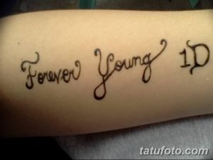 Фото тату с надписью 'Forever Young 1D' на предплечье. Татуировка для фанатов группы One Direction, выполненная в каллиграфическом стиле.