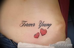 Фото тату с надписью 'Forever Young' и двумя красными сердечками на животе. Нежный рисунок татуировки для девушек, выполненный в ярких цветах.