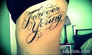 Фото тату на боку: надпись 'Forever Young' выполненная каллиграфическим шрифтом с завитушками и узорами.